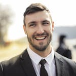 A confident businessman smiling in a black suit with an outdoor background.
