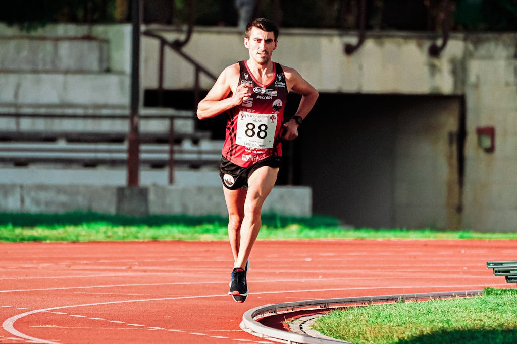 A determined male athlete wearing a bib running on an outdoor track in a stadium setting.