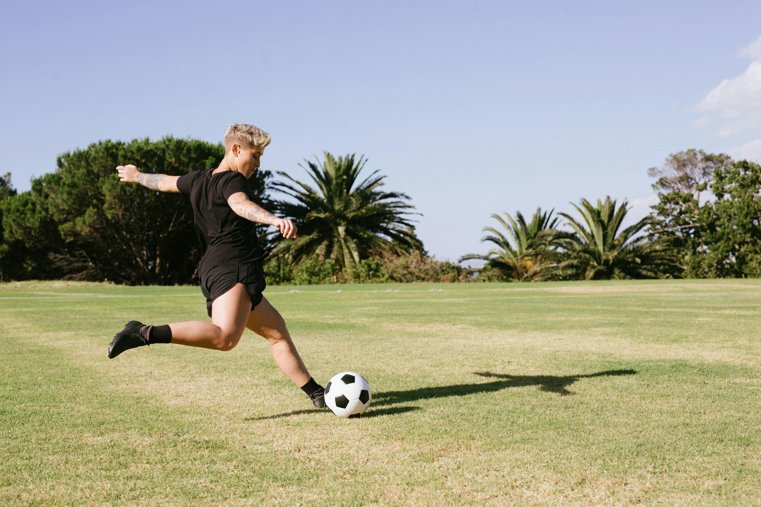 A woman soccer player kicking a ball on a grassy field during a sunny day outdoors.