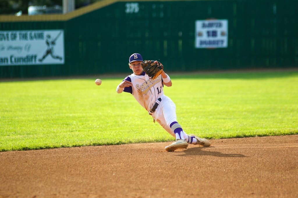 A young baseball player in uniform dives to catch a ball on a well-lit field.