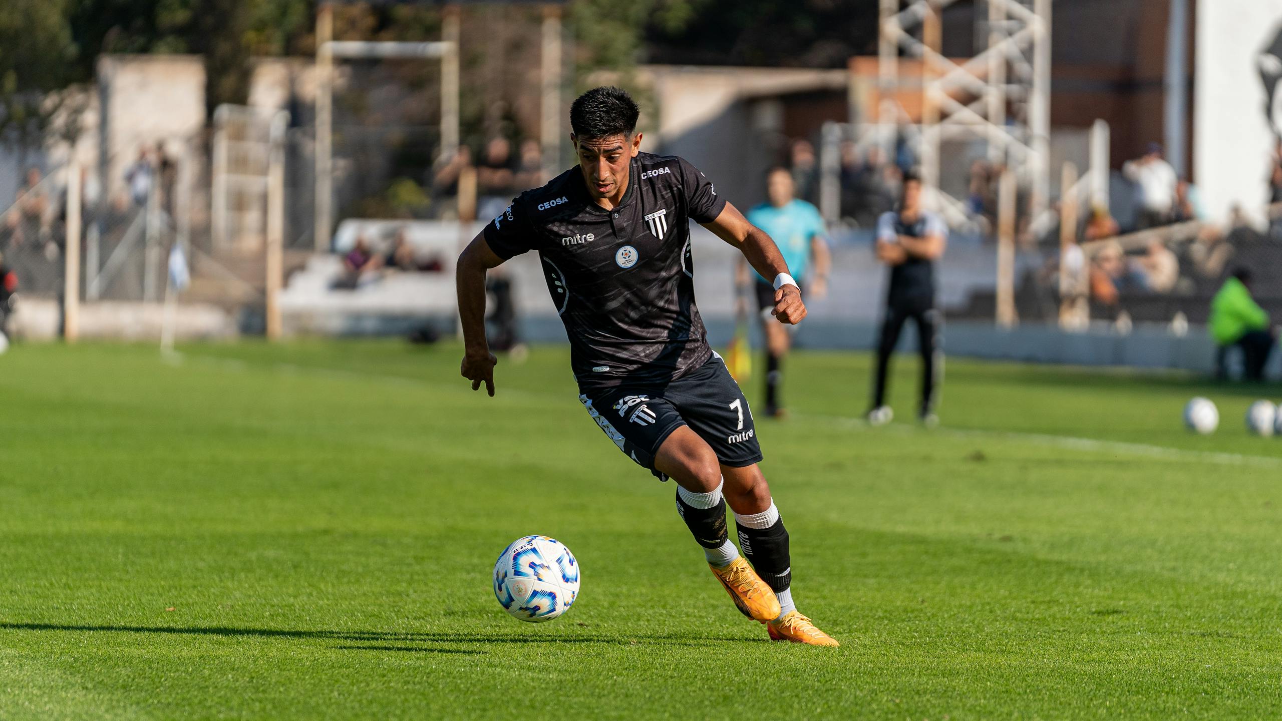 Football player in black jersey dribbles ball on a sunny day match.