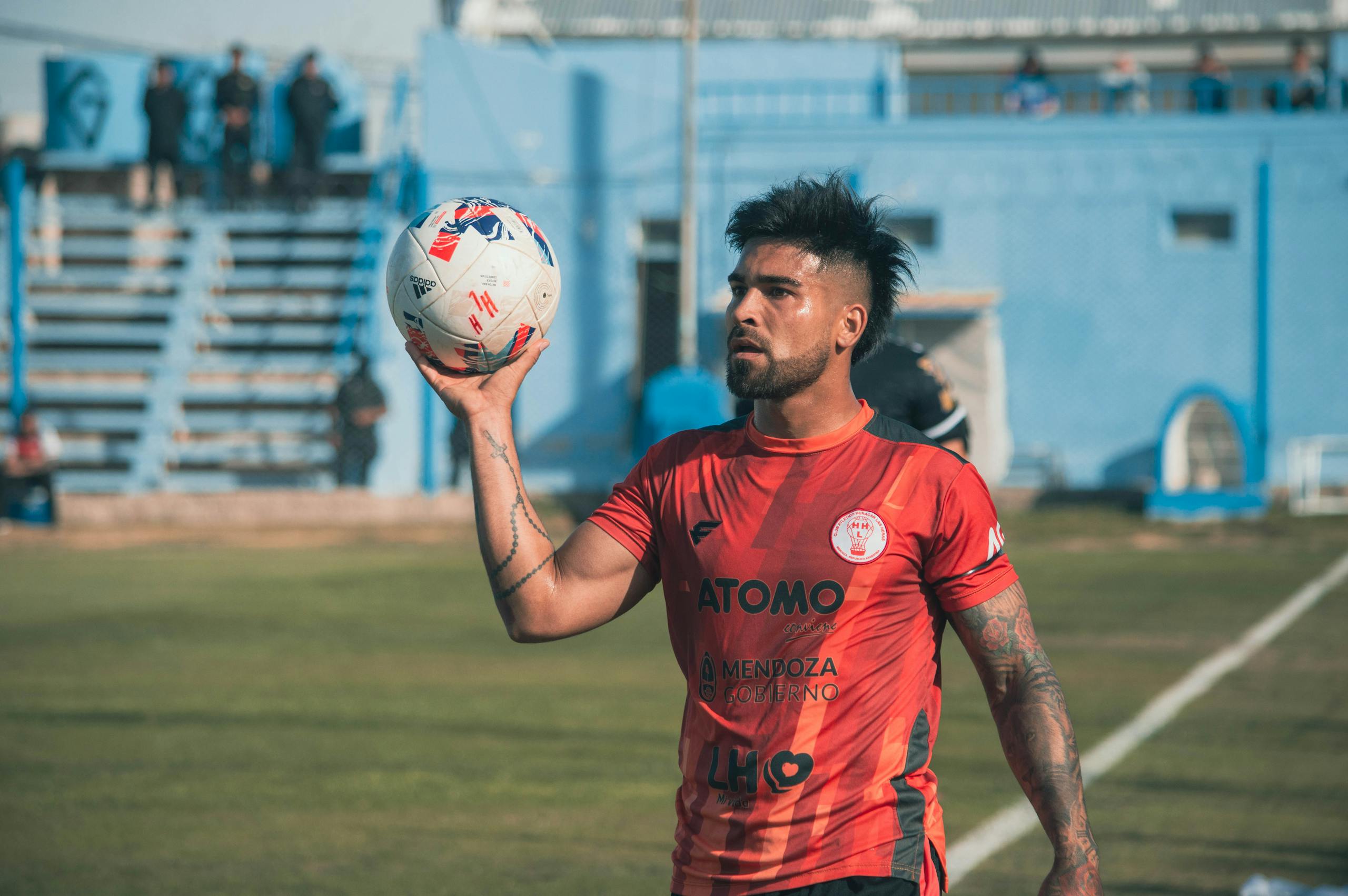 Tattooed soccer player holding a ball, ready for throw-in on a sunny field.