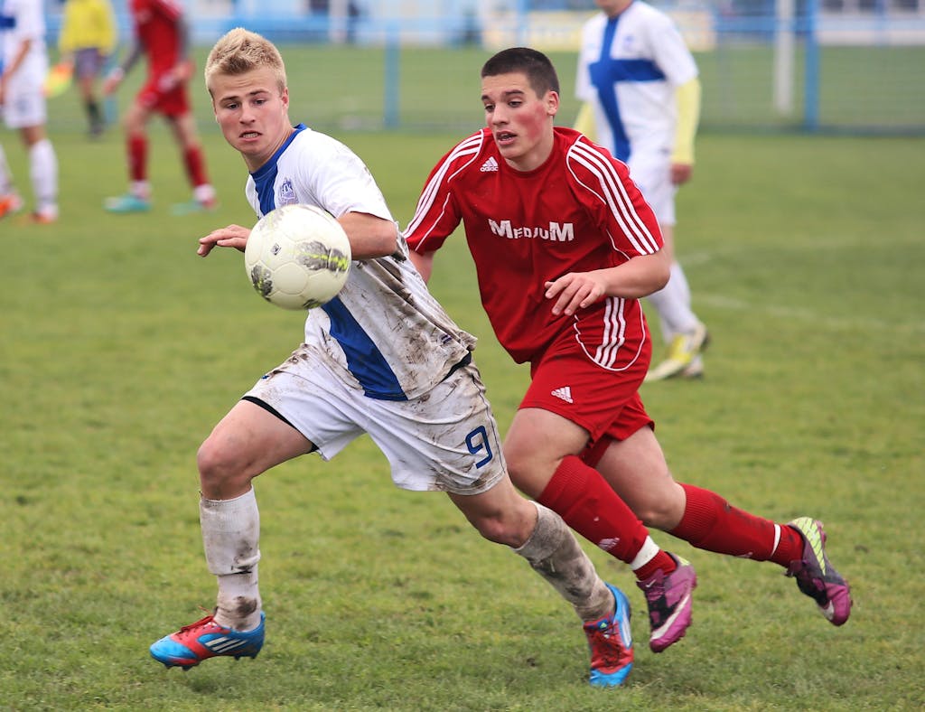 Two teenage soccer players competing fiercely on a muddy grass field.